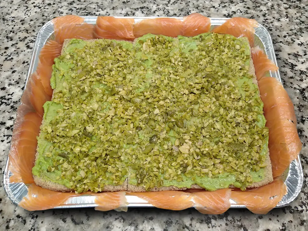 Rebanadas de pan de molde untadas con guacamole y olivas y pepinillos picados.